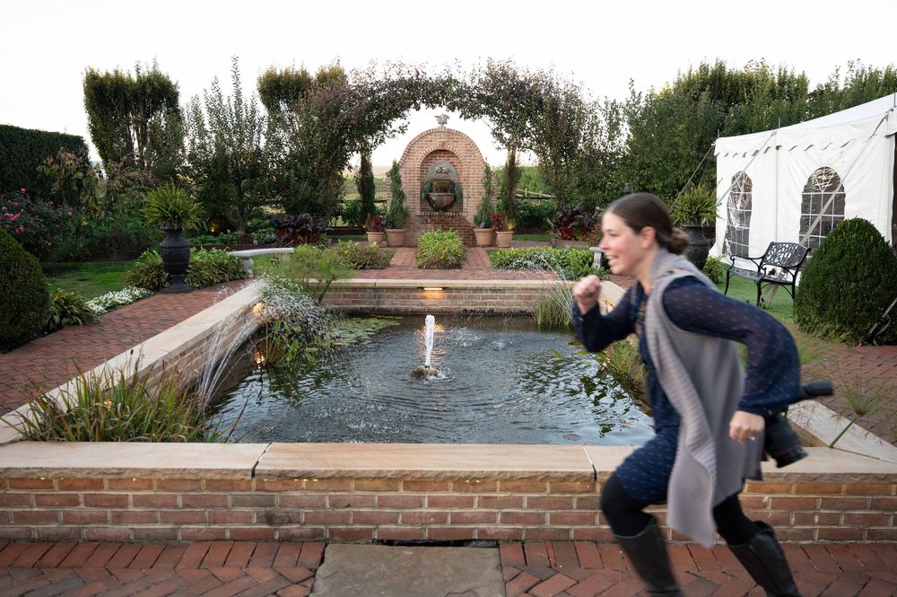 Two people playfully pose by a formal garden fountain with an arched trellis and white tent in the background.