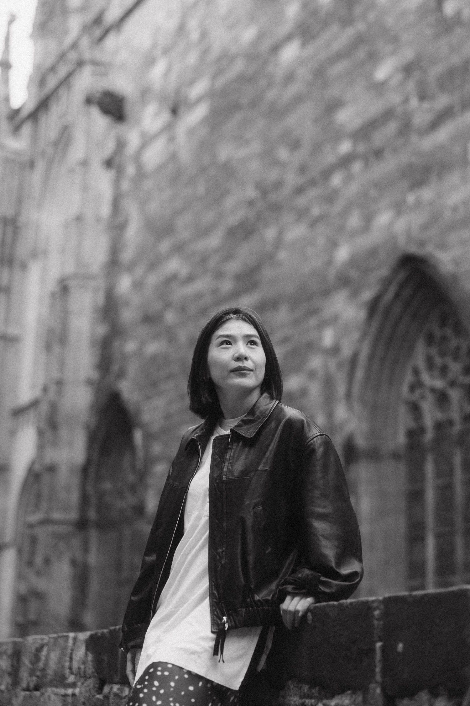Black and white portrait of a young woman in leather jacket leaning against stone wall of Gothic cathedral.