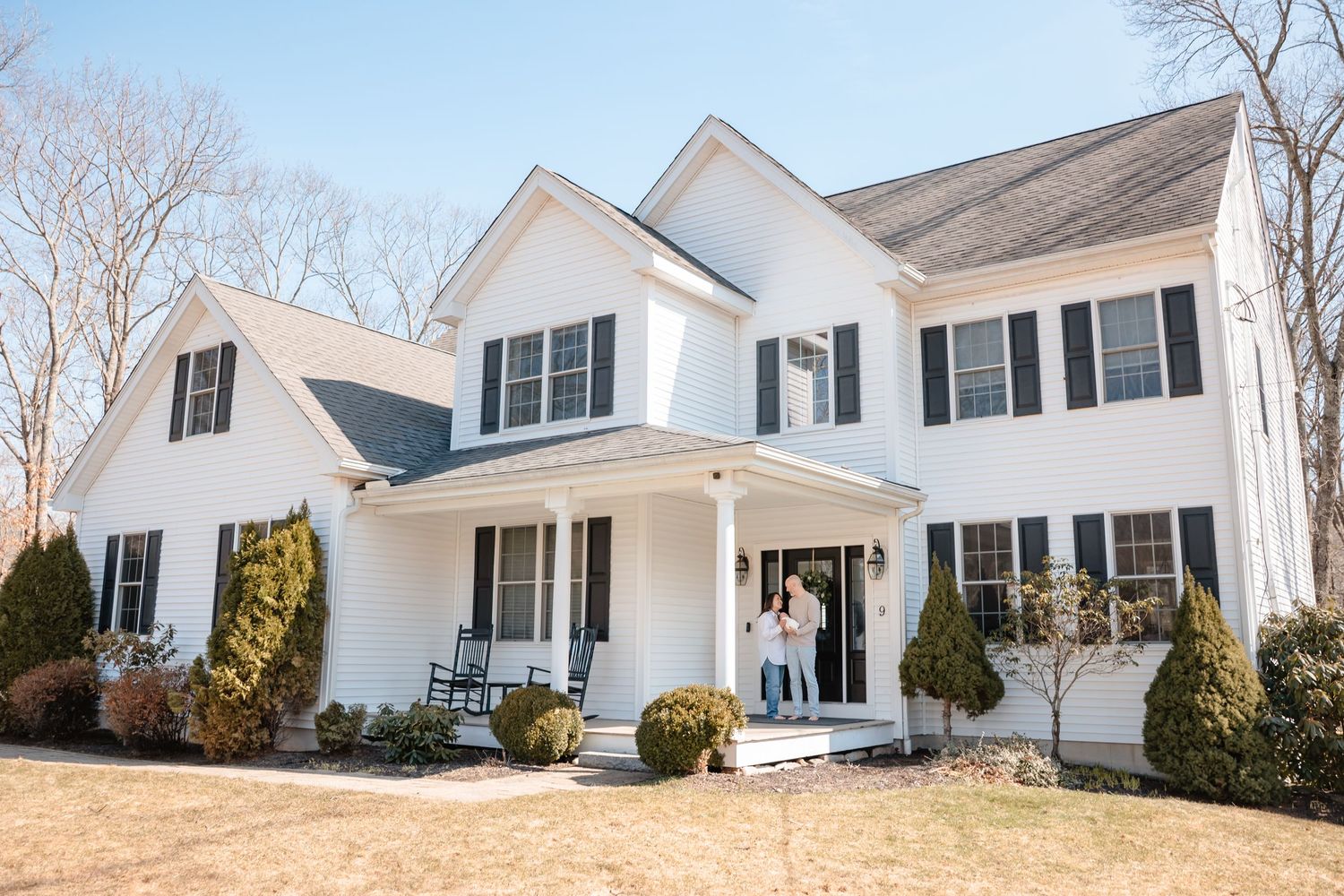 White colonial home with black shutters and covered porch where couple stands, surrounded by mature trees on a sunny day.