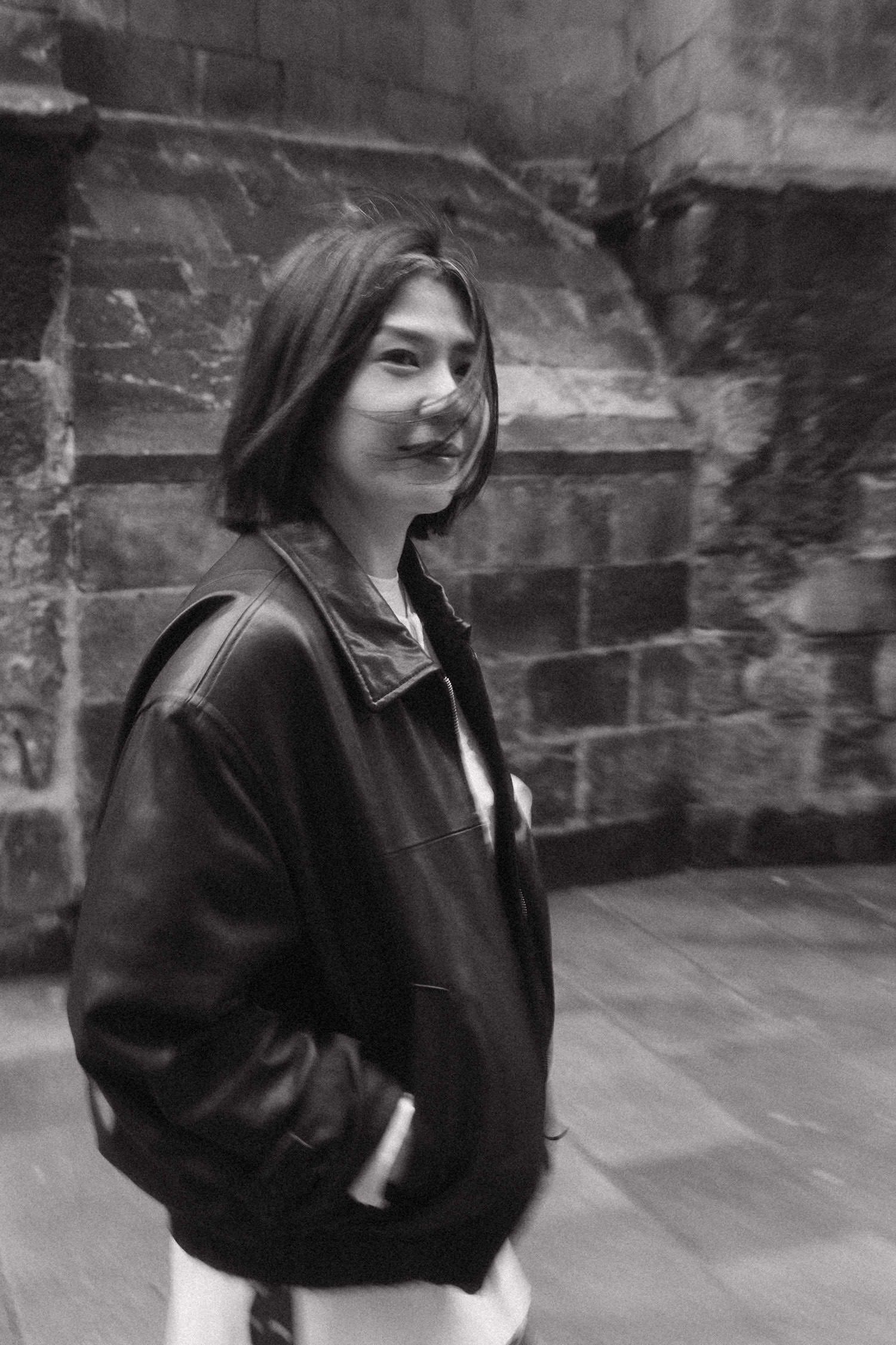 Black and white photo of a smiling young woman in a leather jacket standing against a stone wall.