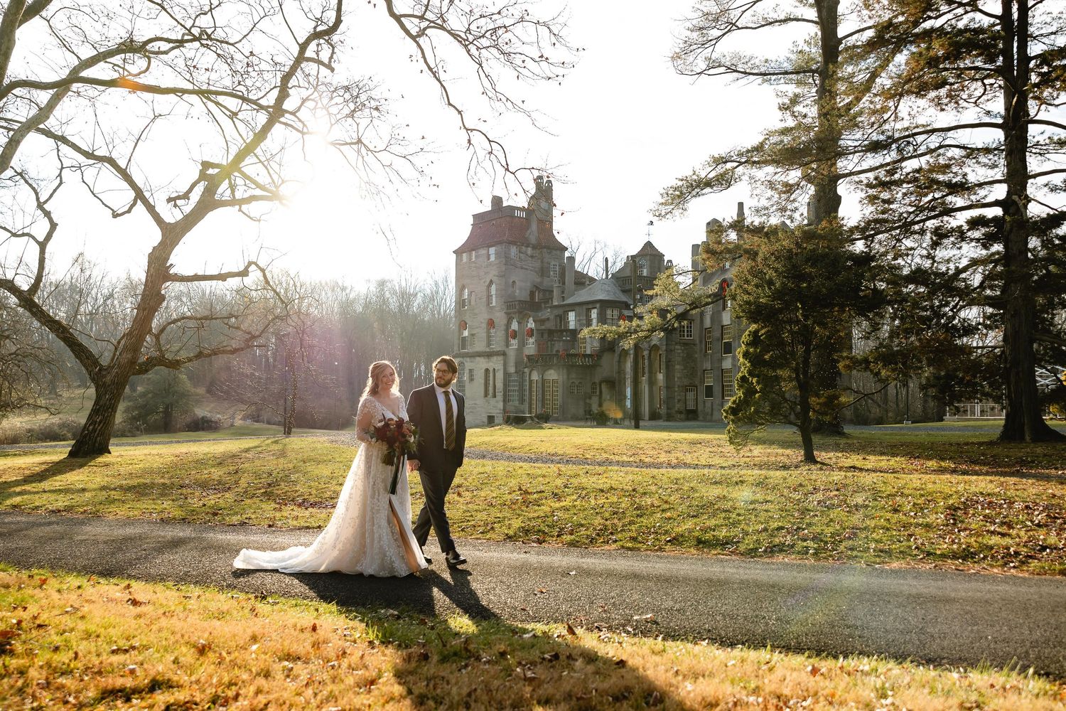 A couple embraces under a sunlit tree in an autumn park setting with golden leaves scattered on the ground.
