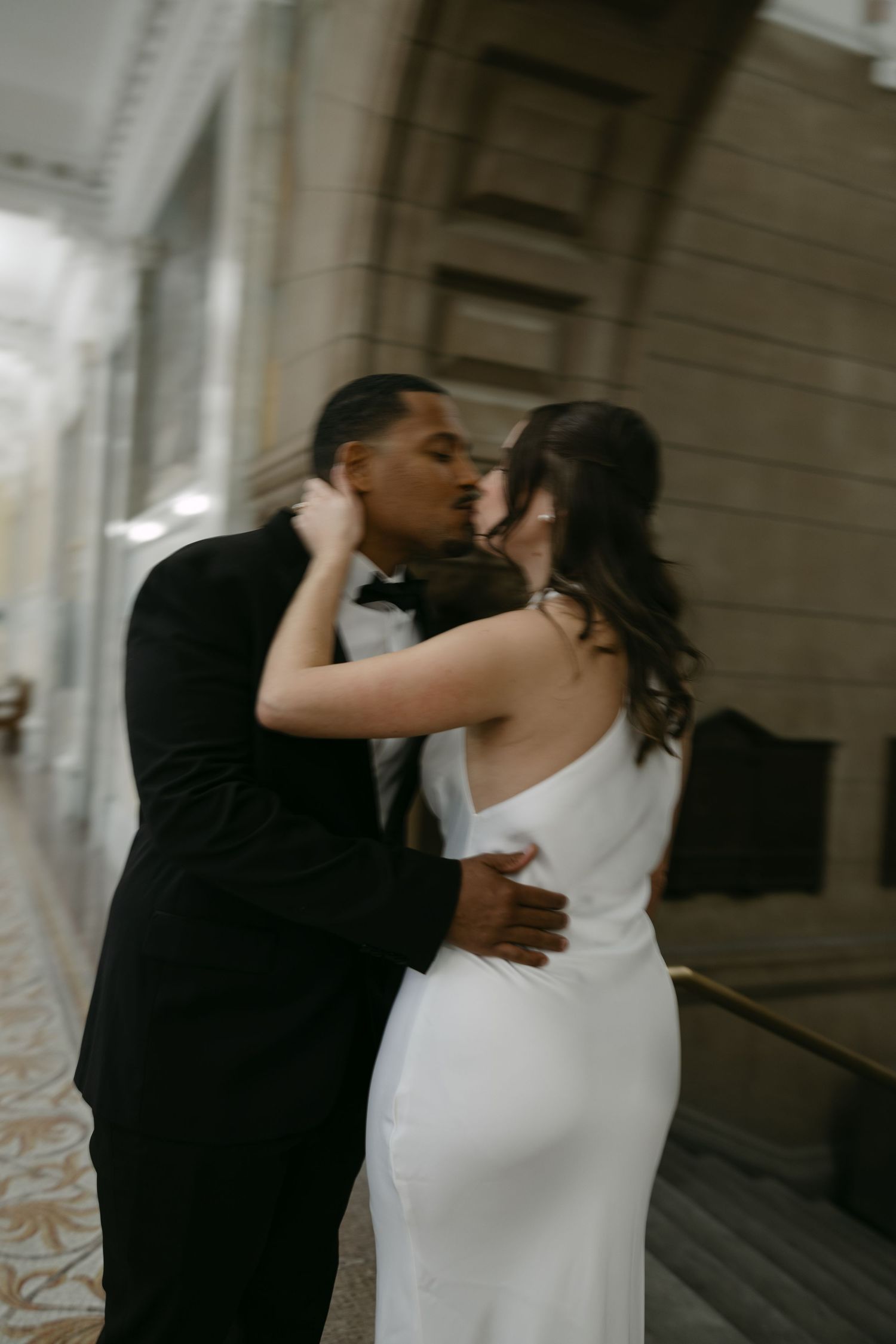 A romantic wedding couple sharing a kiss in formal attire against an elegant architectural backdrop.