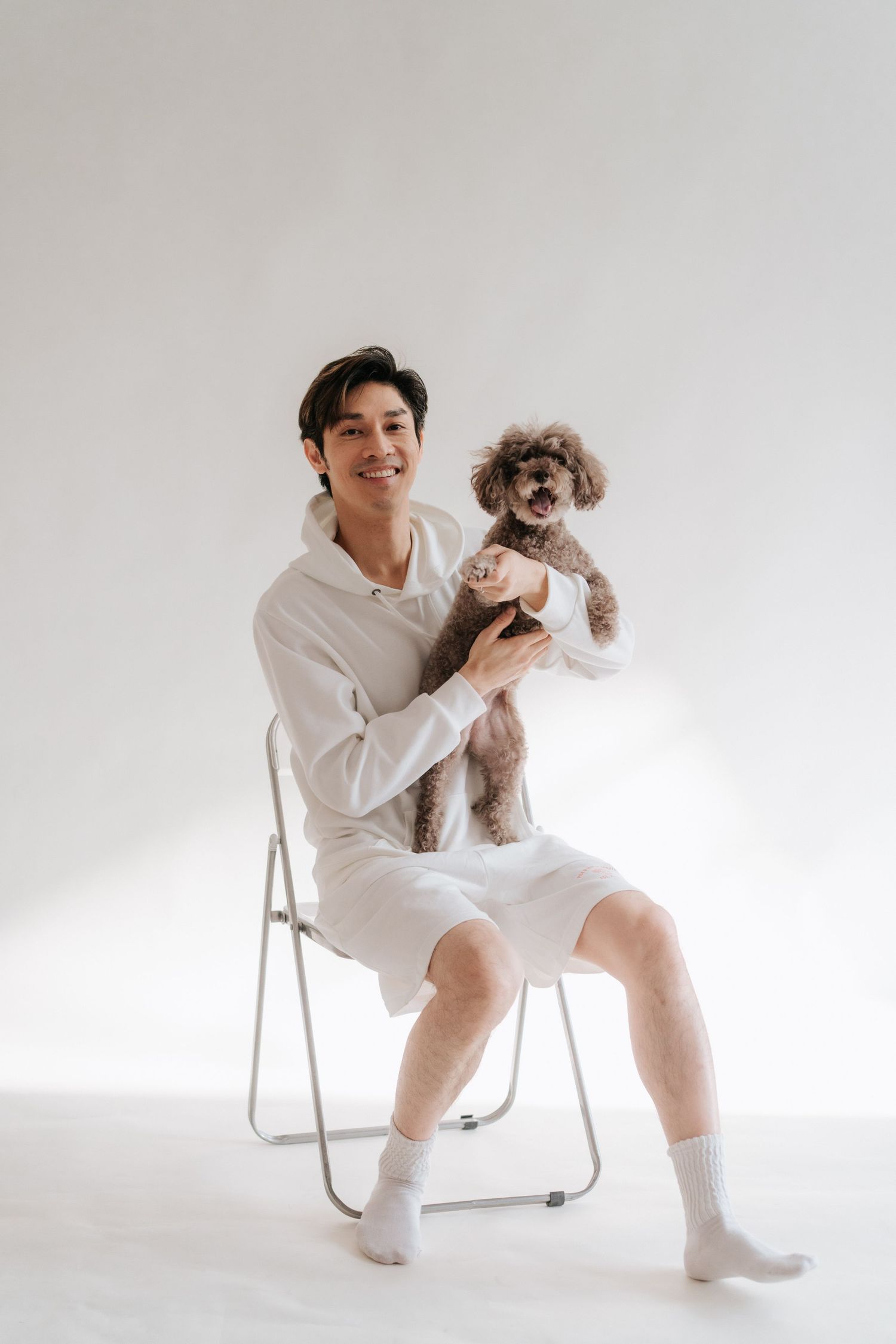 Smiling young man in white outfit sitting on folding chair holding a small gray poodle against white background.一位面帶笑容的年輕男子身穿白色衣服，坐在折疊椅上
