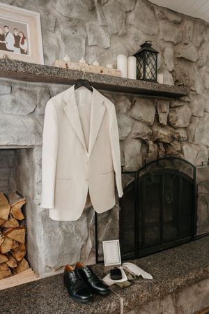 White suit jacket hangs near stone fireplace with black dress shoes and lantern on rustic mantel shelf.