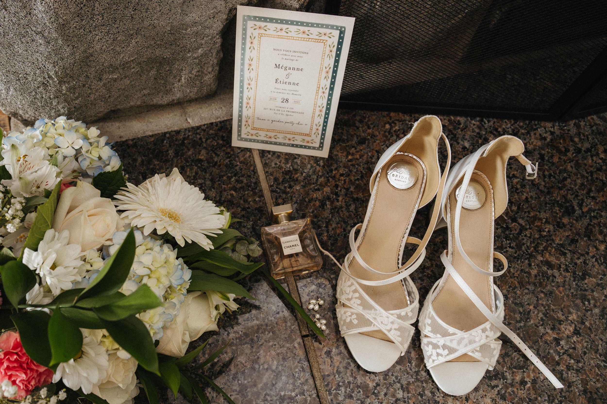 Elegant white bridal shoes displayed next to a framed certificate and delicate floral bouquet with white and pink blooms.