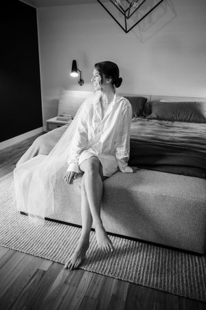 A black and white photograph of someone in a white shirt sitting on a bed in a minimalist bedroom with a wall lamp.