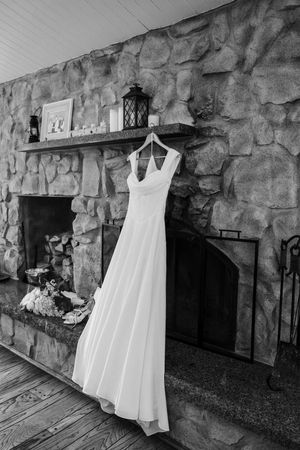 A white wedding dress hangs elegantly against a rustic stone wall fireplace in black and white photography.