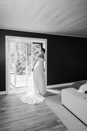 A bride in a flowing white gown stands elegantly by a bright window in a room with dark walls and hardwood floors.