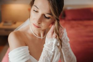A bride in a white wedding dress with lace details and off-shoulder neckline gazes downward in a soft, romantic moment.