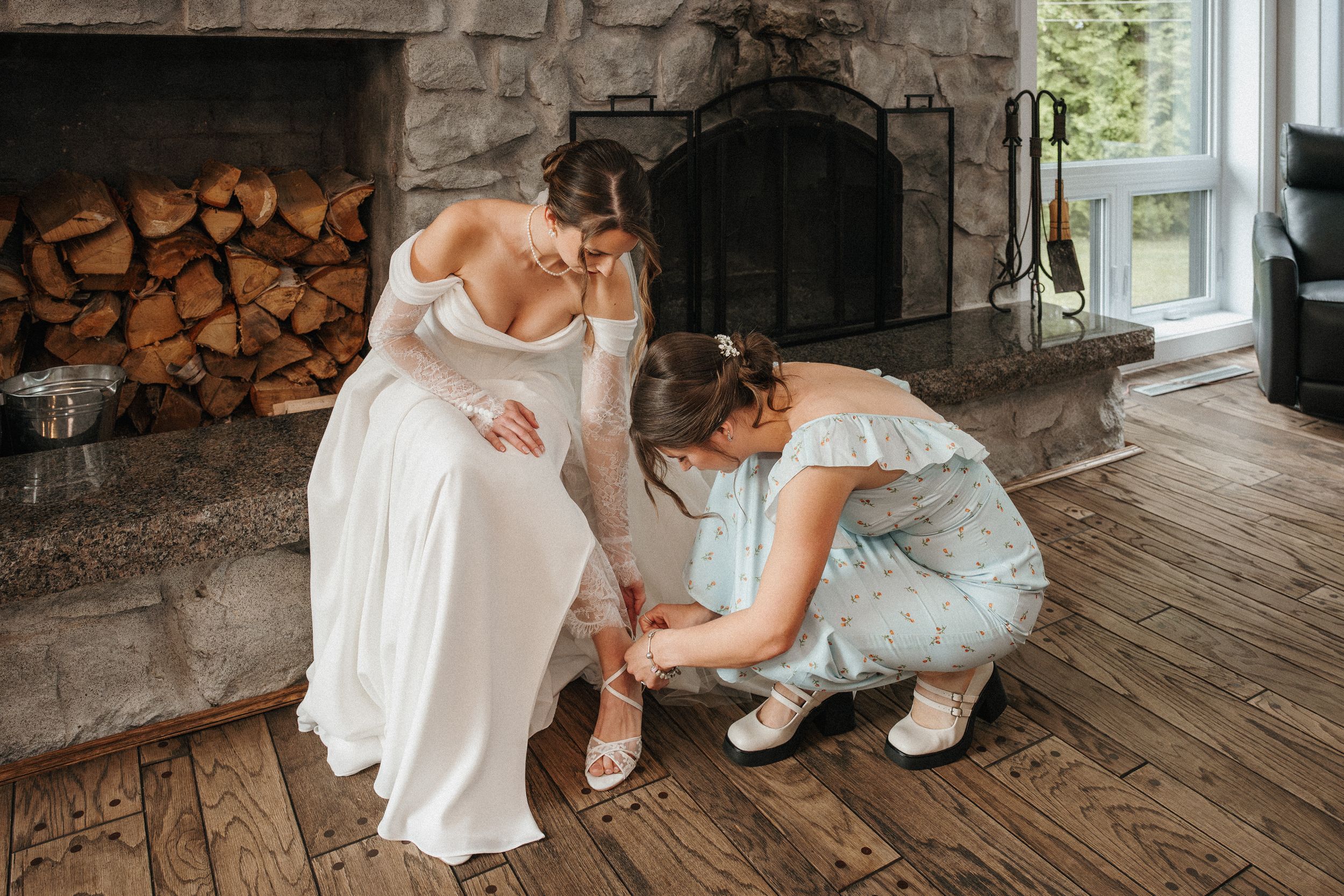 A wedding scene by a stone fireplace with stacked wood shows final dress preparations on hardwood floors.
