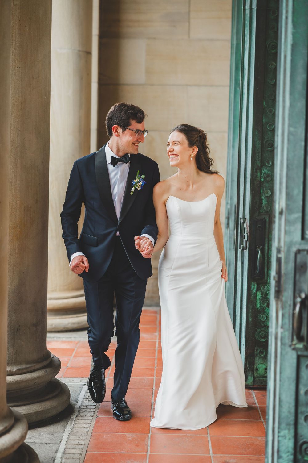A happy couple in wedding attire walks together and laughs along a covered outdoor walkway with columns and terracotta tiles.