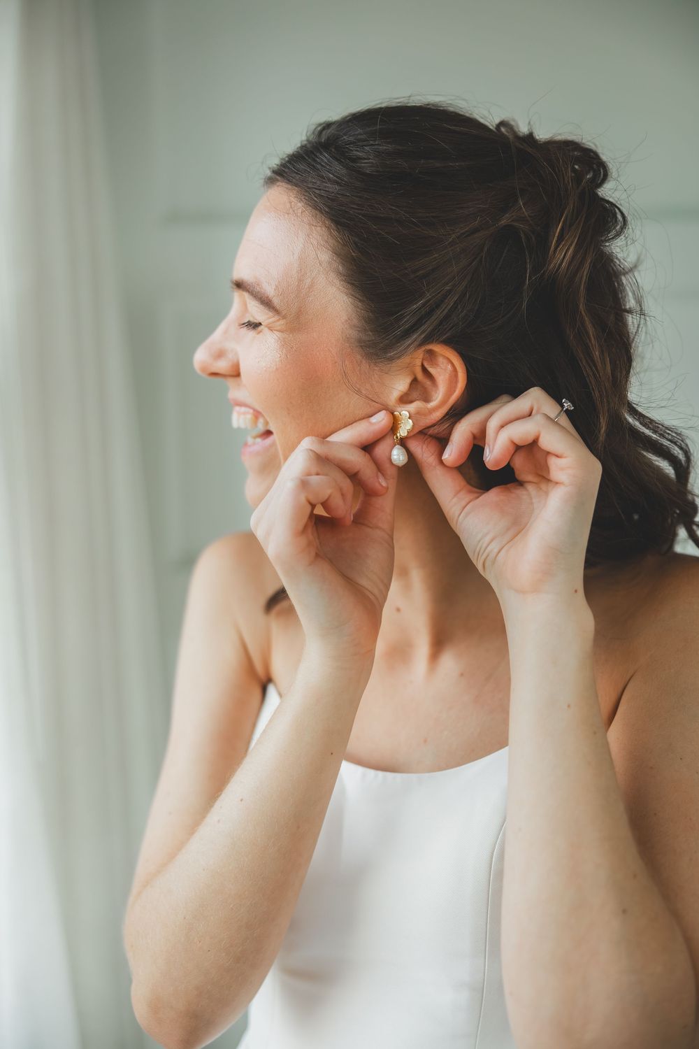 Side profile of a bride wearing a strapless gown putting on earrings while smiling with their hair in an updo.