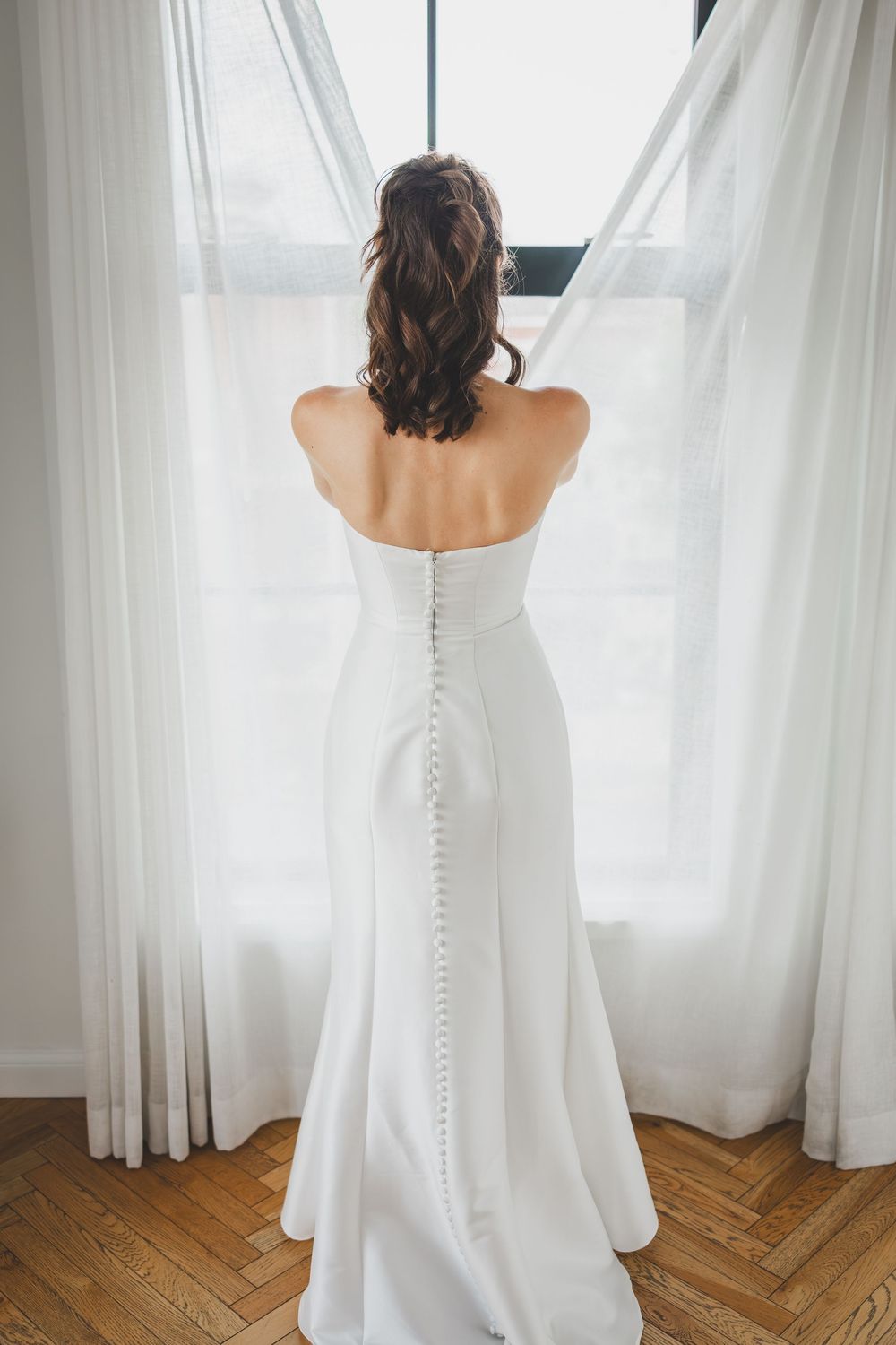 Back view of bride in elegant white strapless wedding gown with button details standing by sheer curtained window.