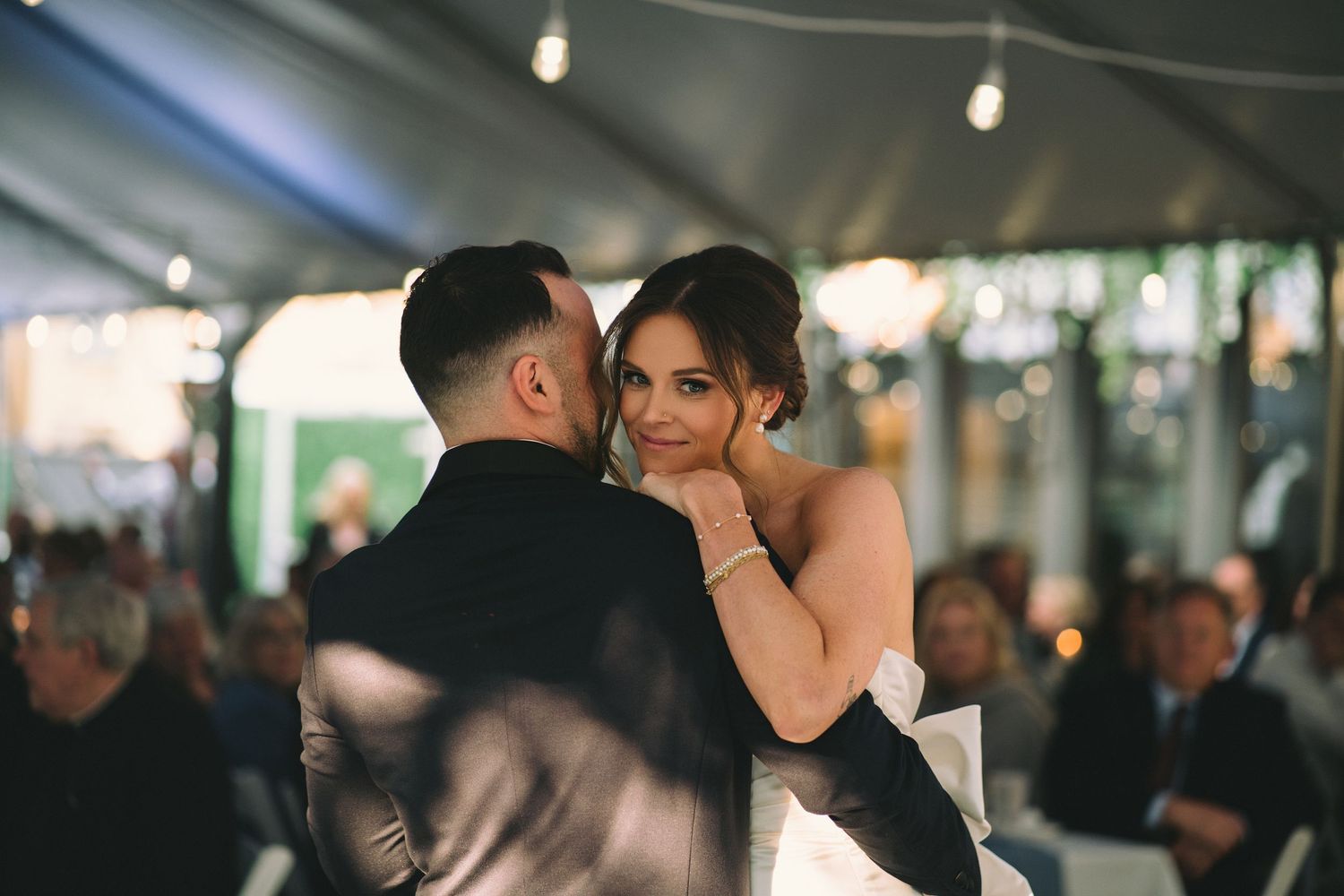 A couple shares a romantic dance moment at a dimly lit wedding reception with other guests in the background.