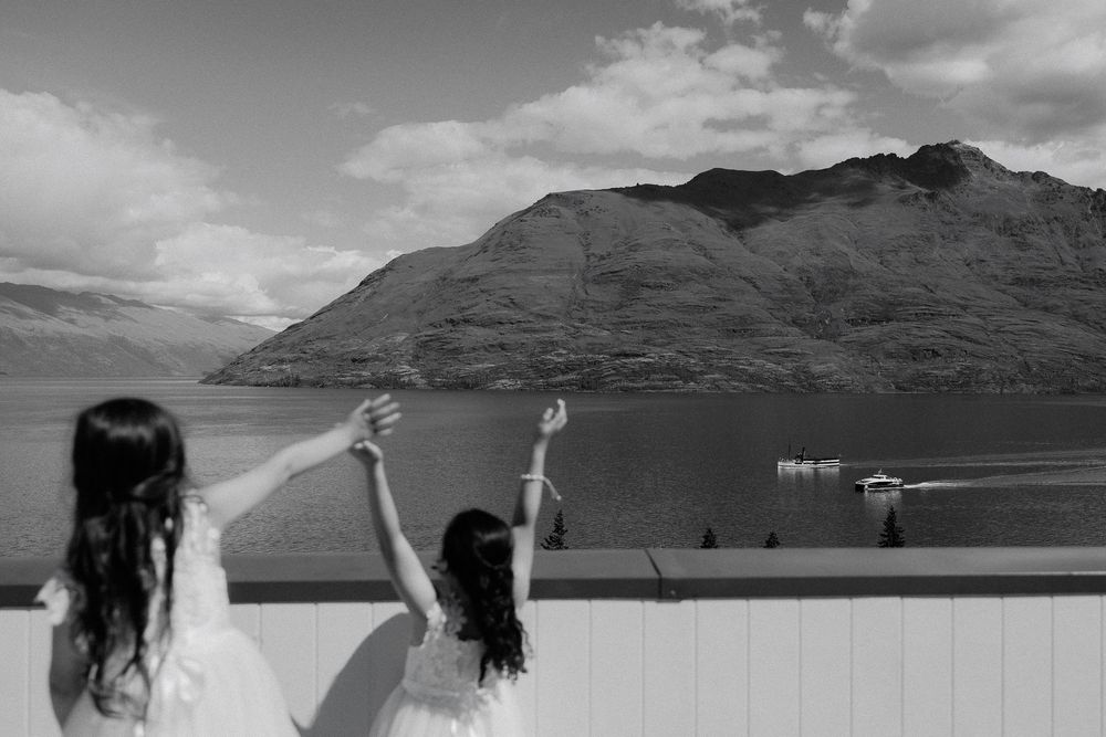 Two flower girls in white dresses look out over Lake Wakatipu from Rendezvous Heritage Hotel, Queenstown wedding venue, New Zealand.