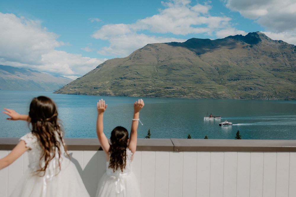 Two flower girls in white dresses look out over Lake Wakatipu from Rendezvous Heritage Hotel, Queenstown wedding venue, New Zealand.
