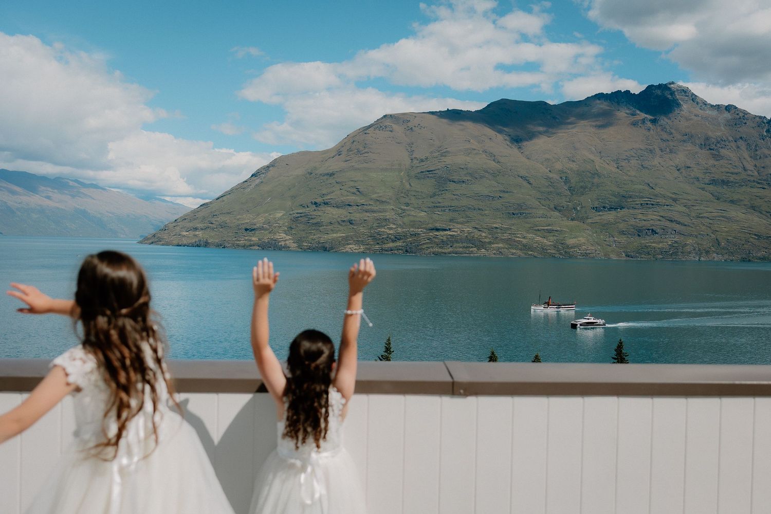 Two flower girls in white dresses look out over Lake Wakatipu from Rendezvous Heritage Hotel, Queenstown wedding venue, New Zealand.