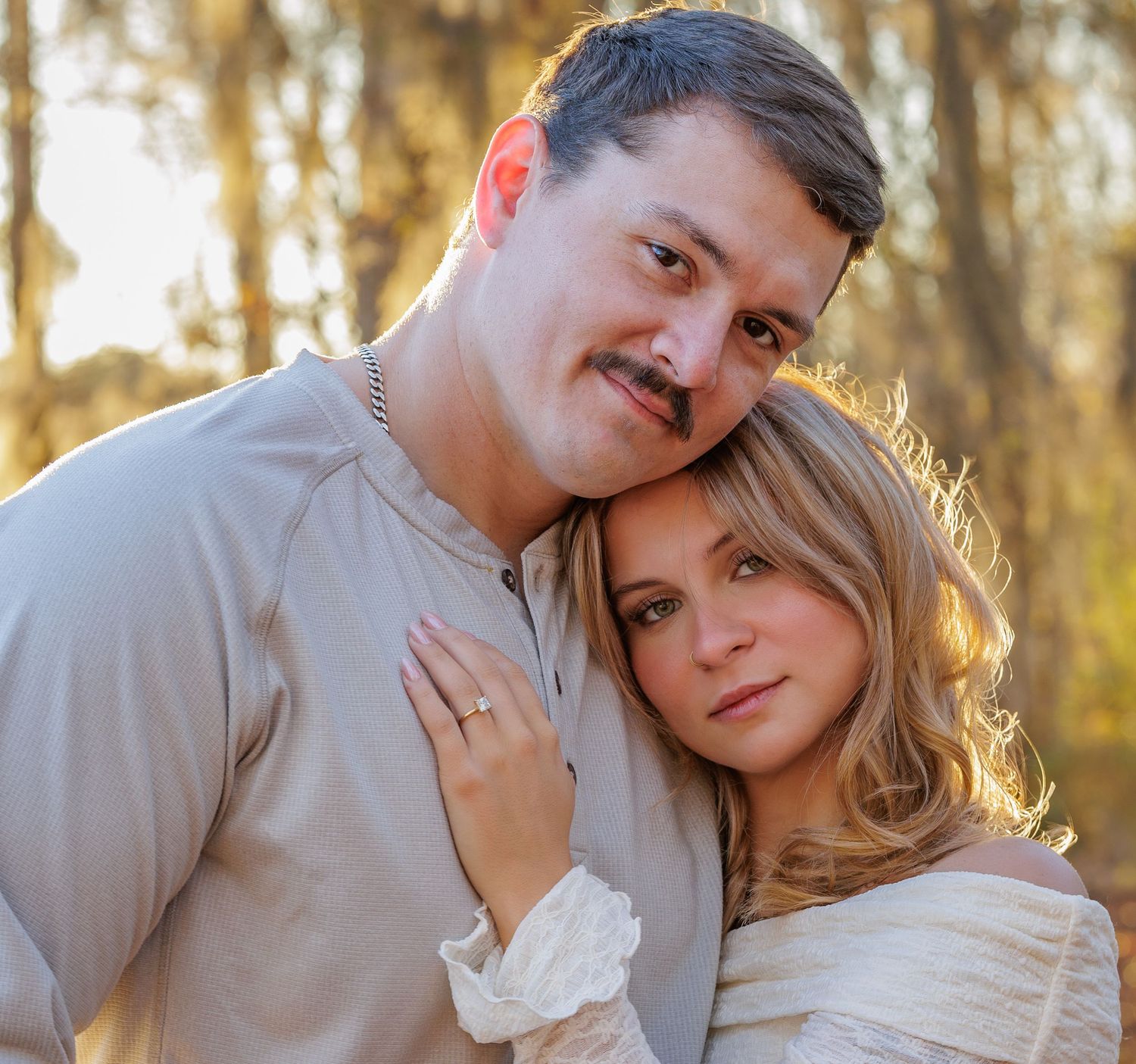 A romantic outdoor portrait of a couple in light colored clothing with golden sunlight filtering through trees behind them.