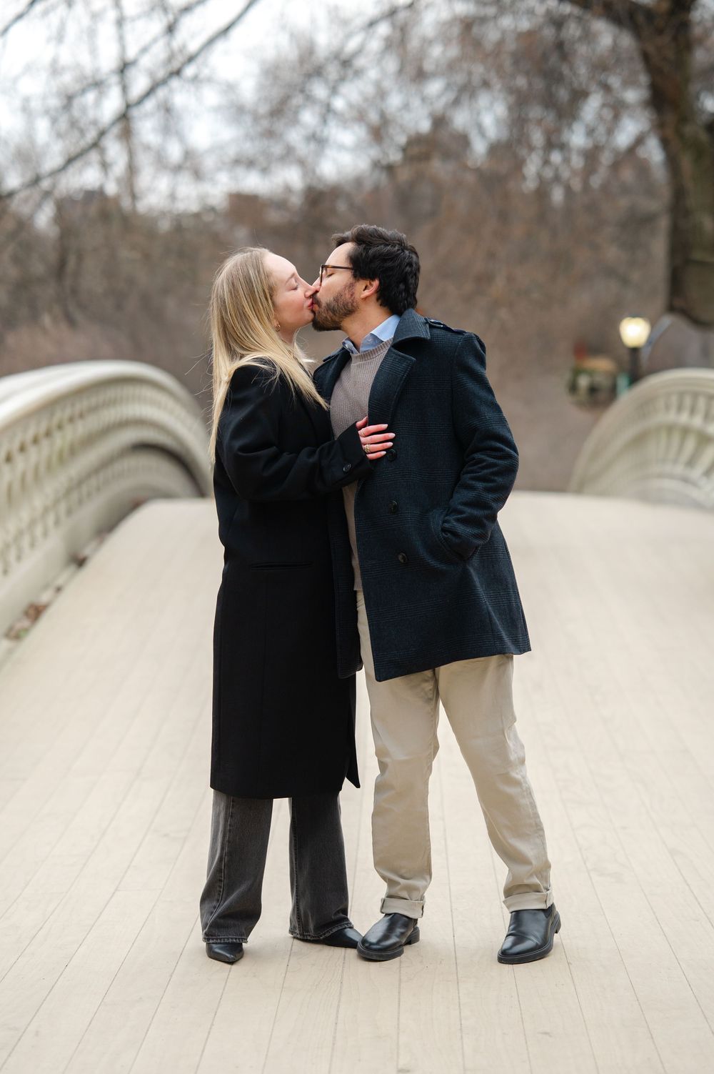 A romantic couple shares a tender kiss while standing on a white bridge in a park during winter.