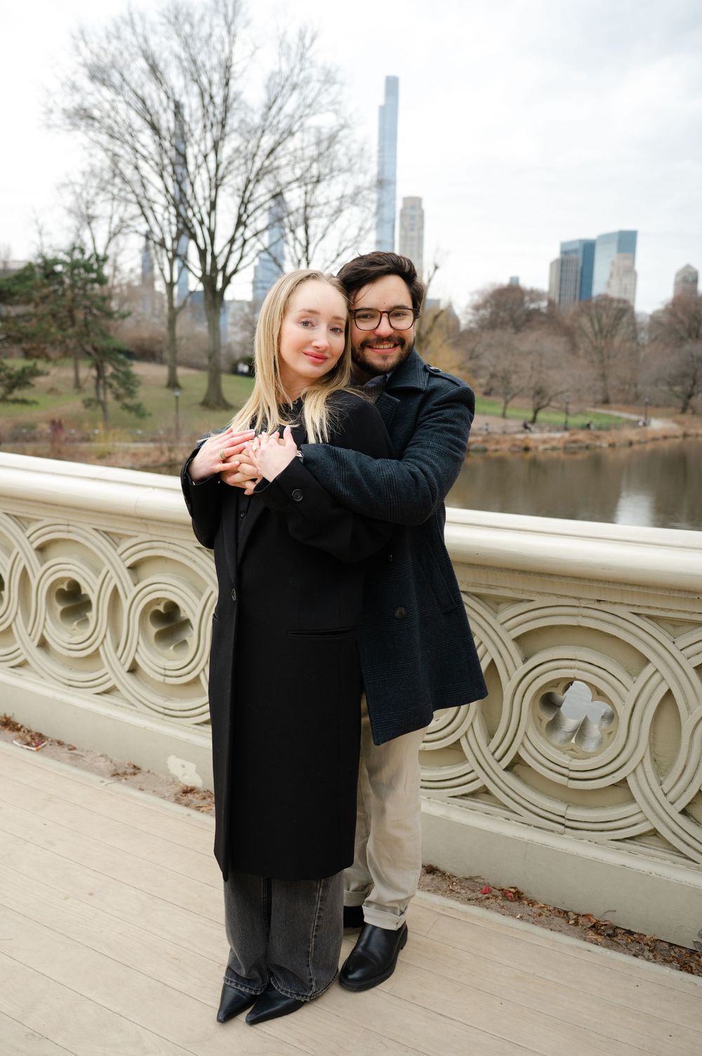A couple embraces on Bow Bridge in Central Park with Manhattan skyscrapers visible in the background on a winter day.