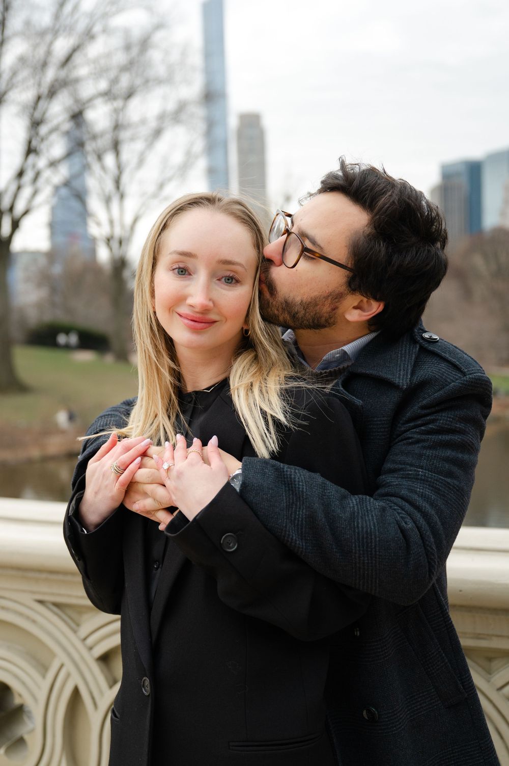 A couple shares a romantic moment in Central Park with the NYC skyline visible in the background.