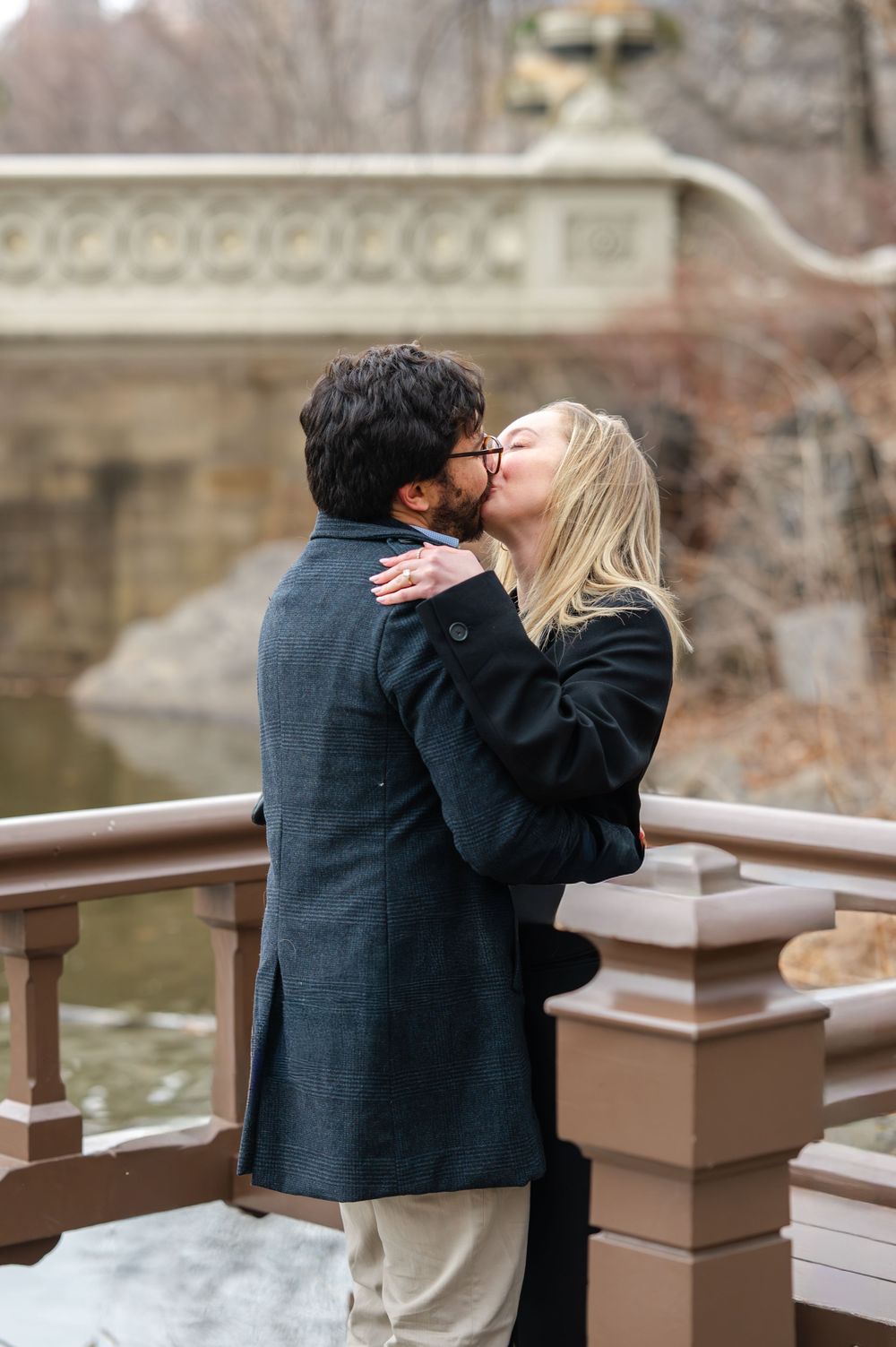 A couple shares a romantic kiss while standing at a wooden railing overlooking water on a winter day.