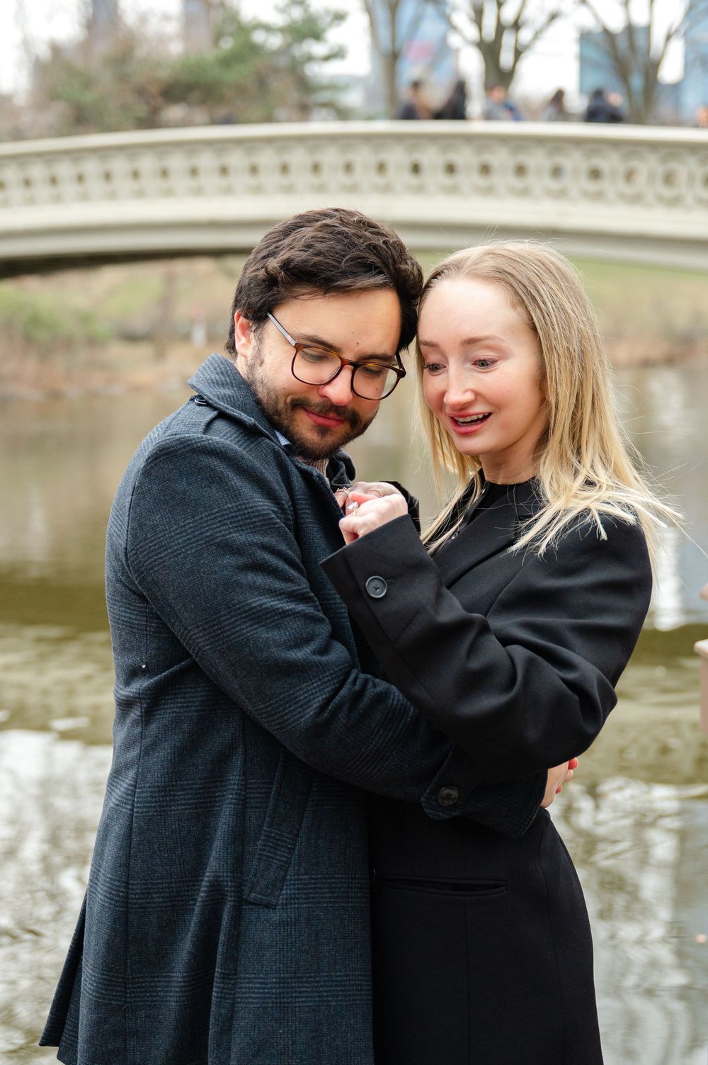 A couple in dark winter coats embraces lovingly by a pond on a cold day, sharing intimate romantic moments together.