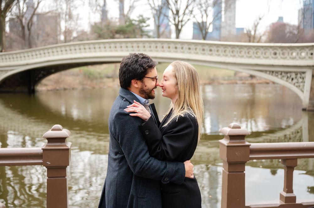 A romantic couple embraces by a stone bridge in Central Park on a winter afternoon.