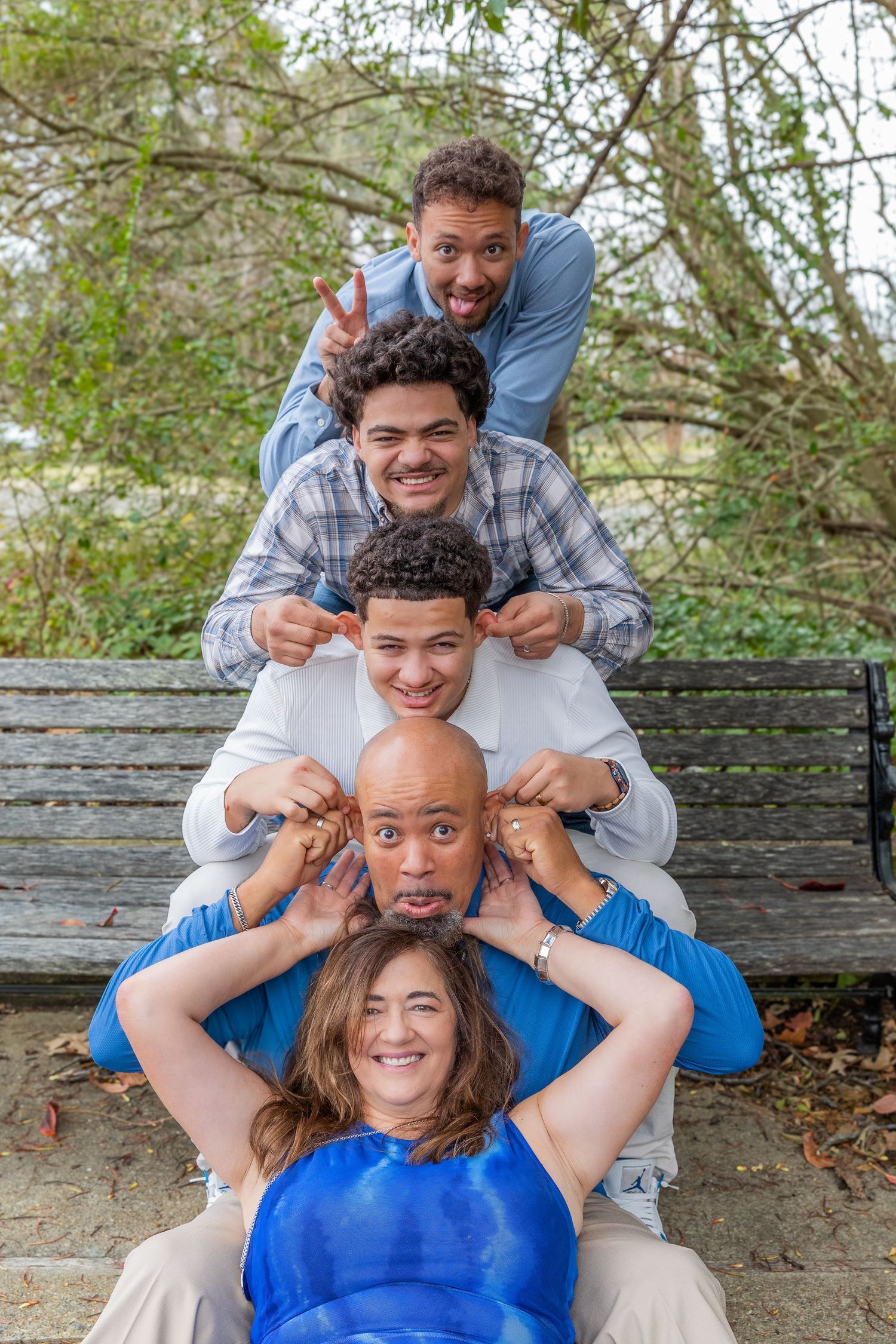 A fun family of five stacks their heads in a totem pole pose on a park bench, laughing and making faces.