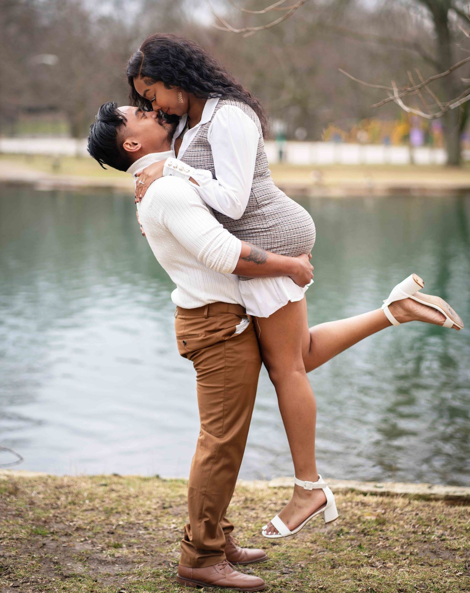 A romantic couple embraces by a lake, with one person lifted up in a playful pose against a serene natural backdrop.