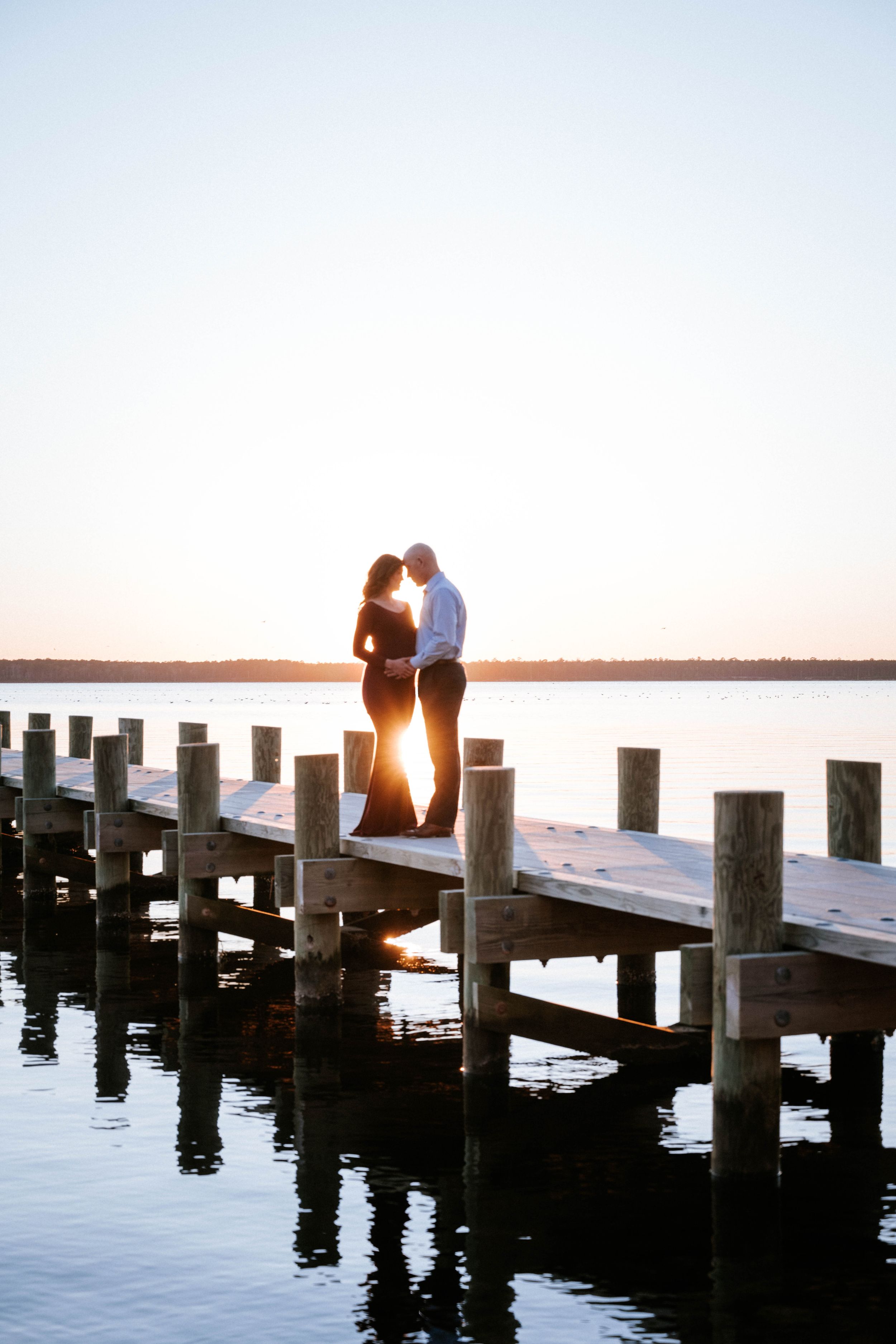 Silhouetted couple sits on wooden pier at sunset with sun glowing between them over calm waters.