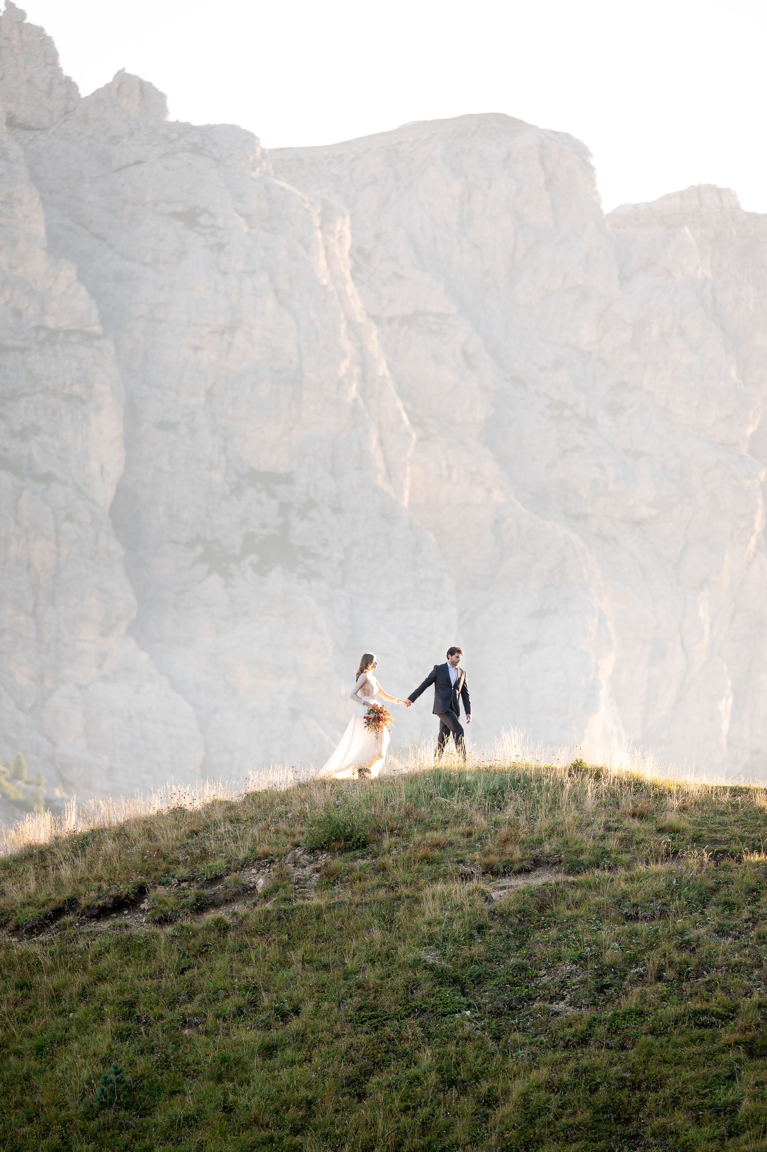 A couple stands atop a grassy hill against towering limestone cliffs in the Dolomites during a mountain wedding.