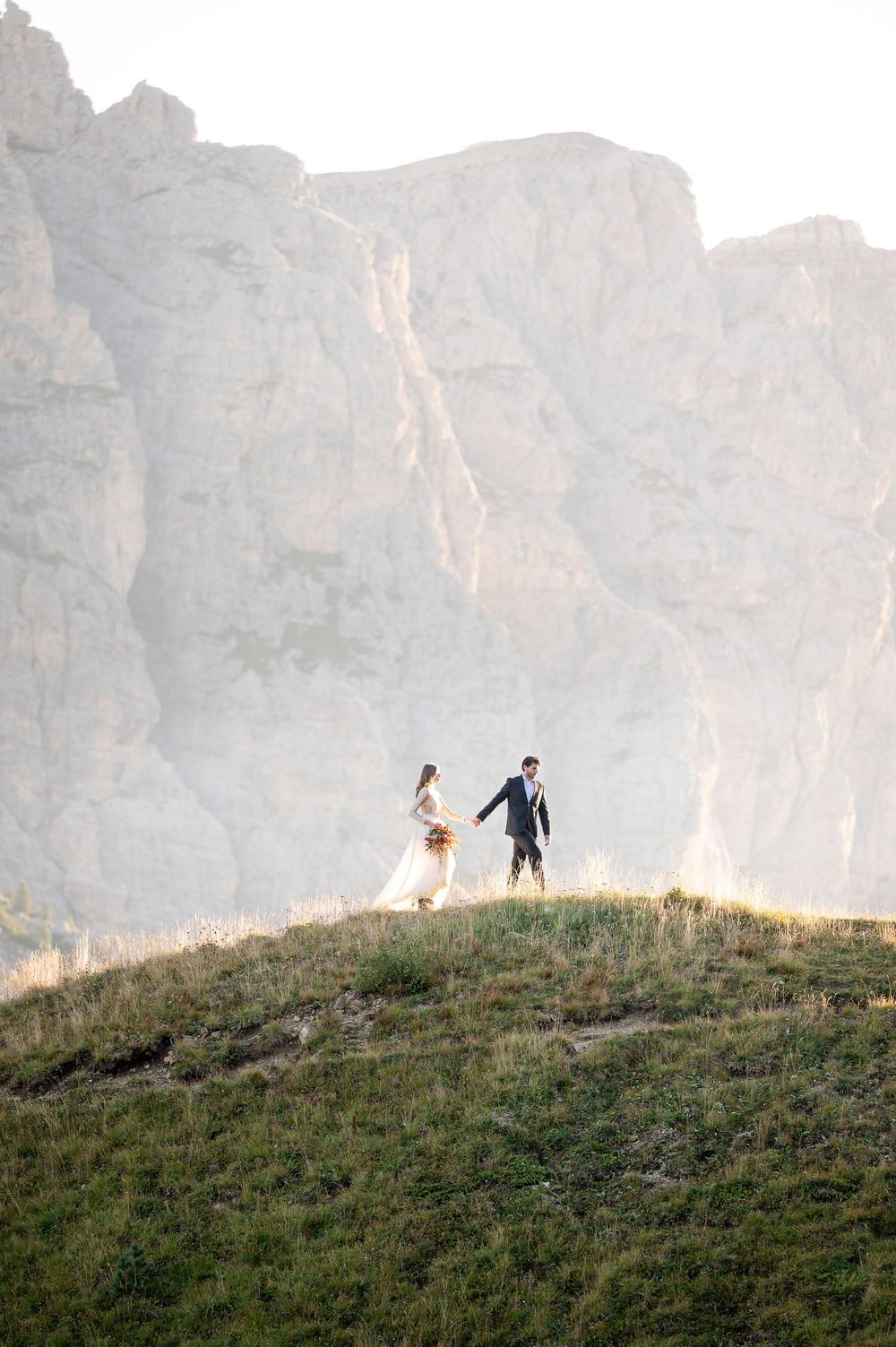A couple stands atop a grassy hill against towering limestone cliffs in the Dolomites during a mountain wedding.