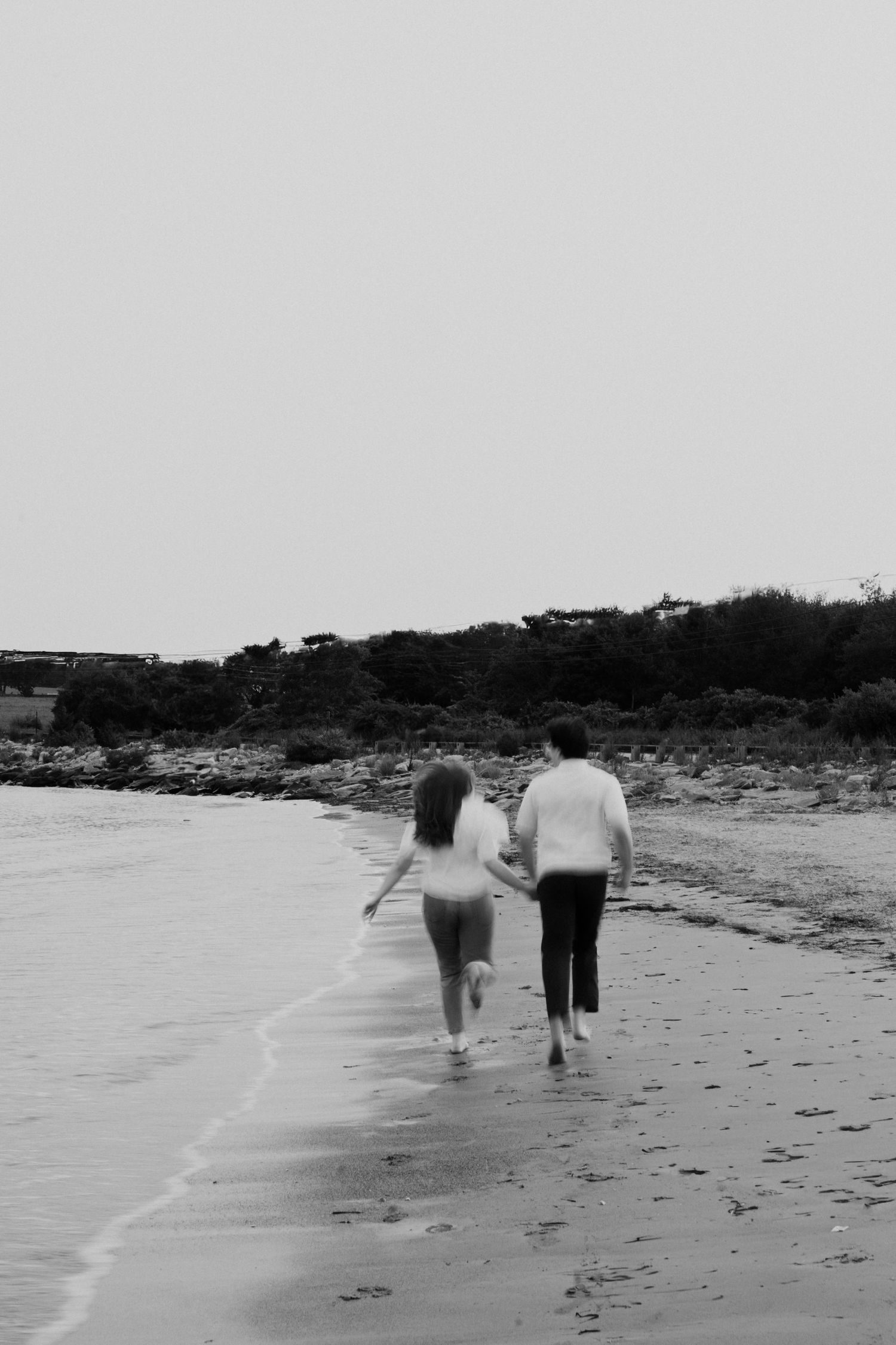 Couple walks hand in hand along beach shoreline in black and white.