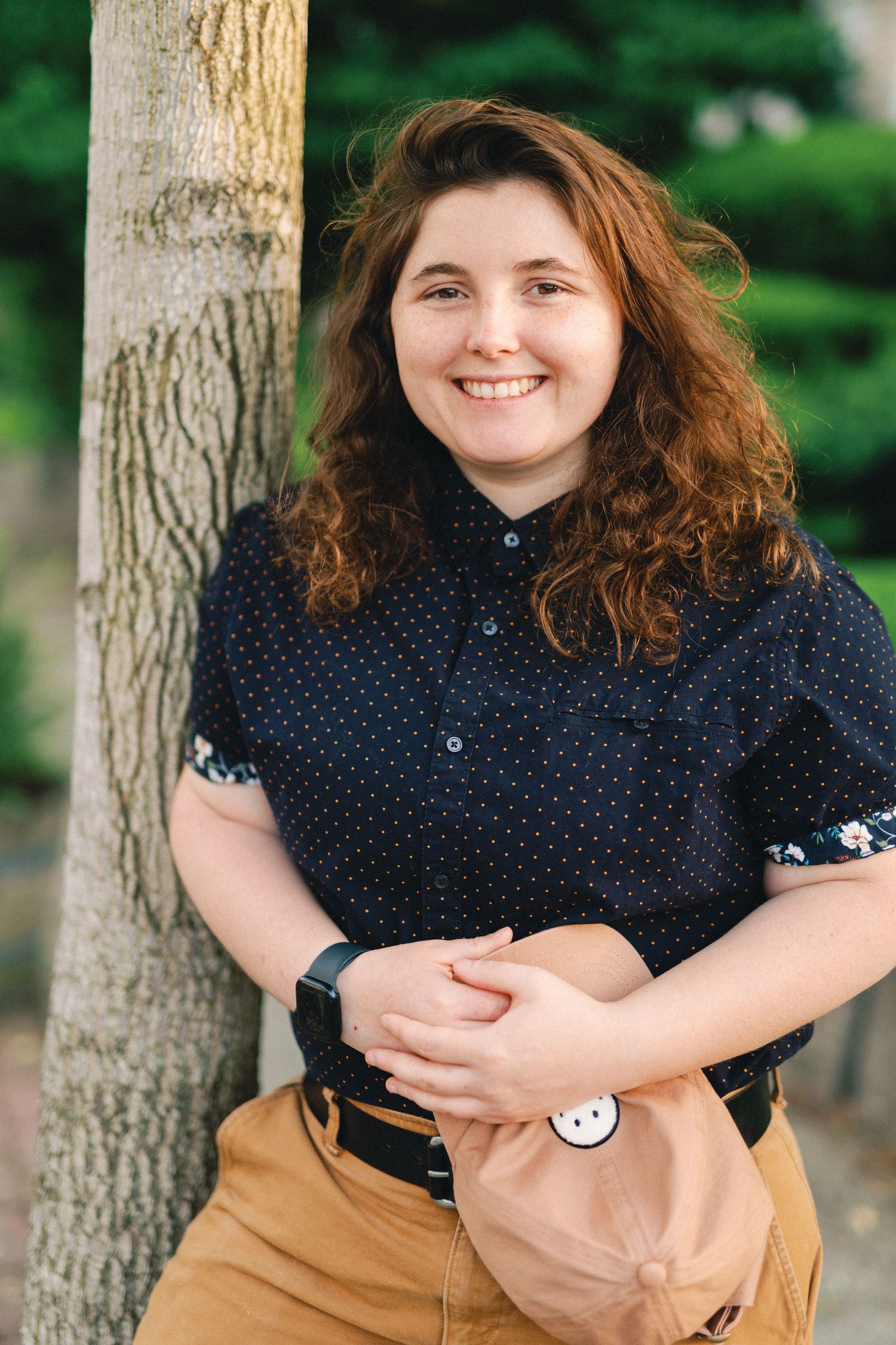 Person with curly brown hair wearing a polka dot navy shirt and khaki pants poses casually against a tree trunk outdoors.