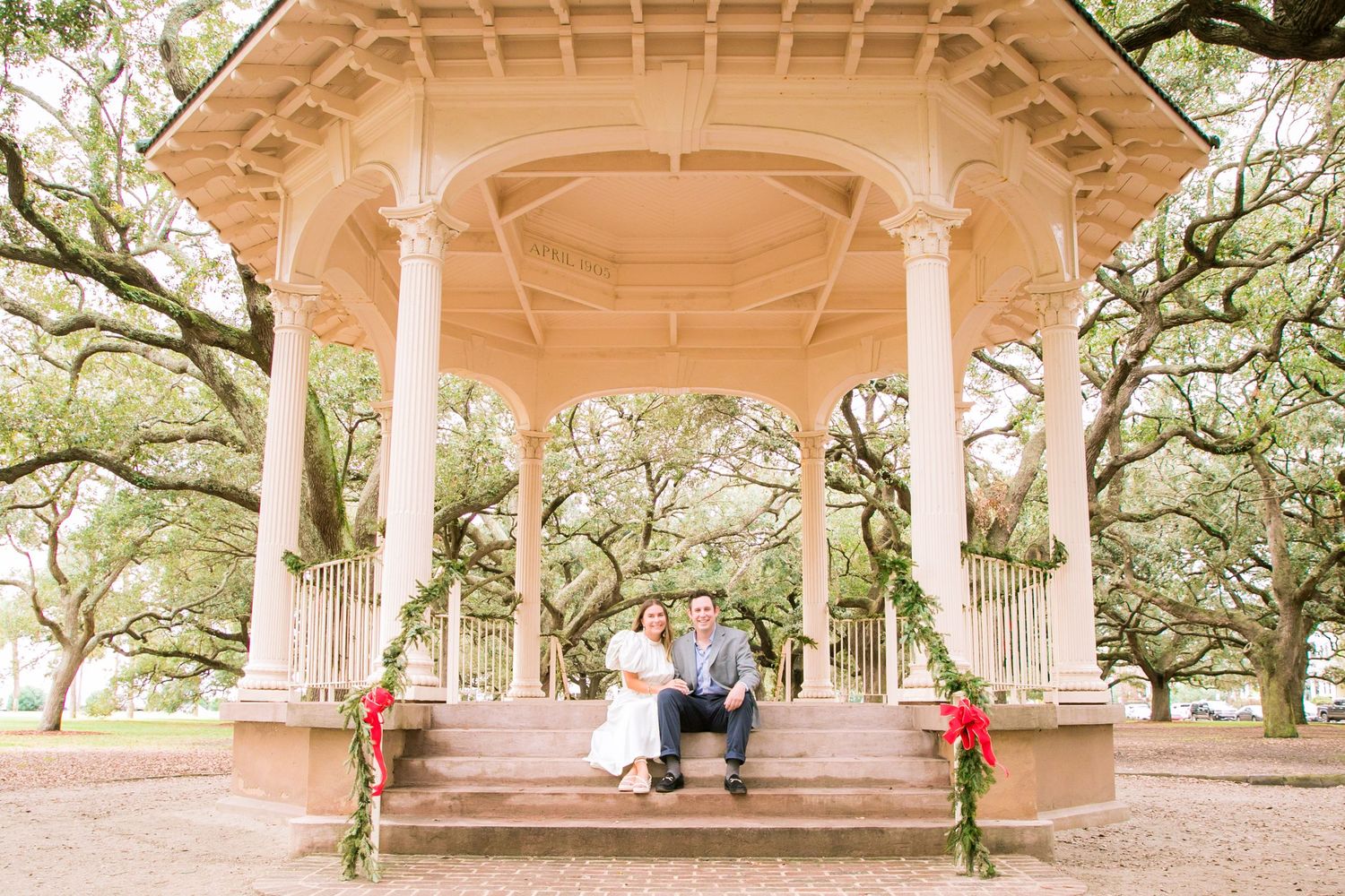 A romantic scene in an ornate white gazebo surrounded by Spanish moss draped oak trees in a park.