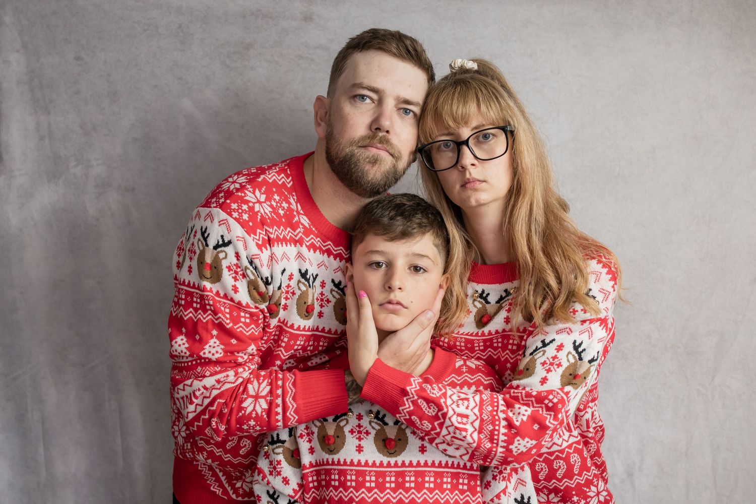 Family of three in matching red Christmas sweaters posing against gray wall.