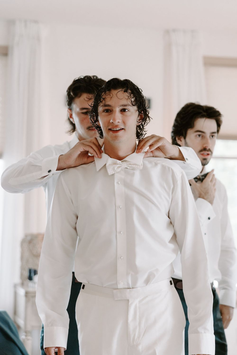 Groomsman ties a white bow tie on the groom while another man adjusts his own tie behind them.