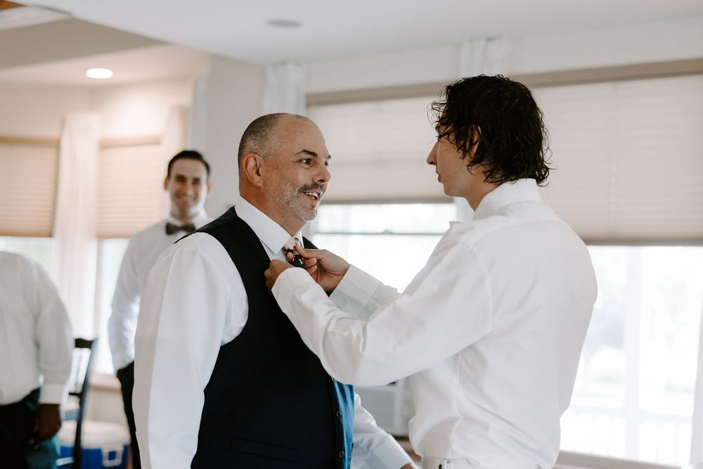 Groom adjusts bow tie on older man in black vest while groomsmen prepare in bright room.