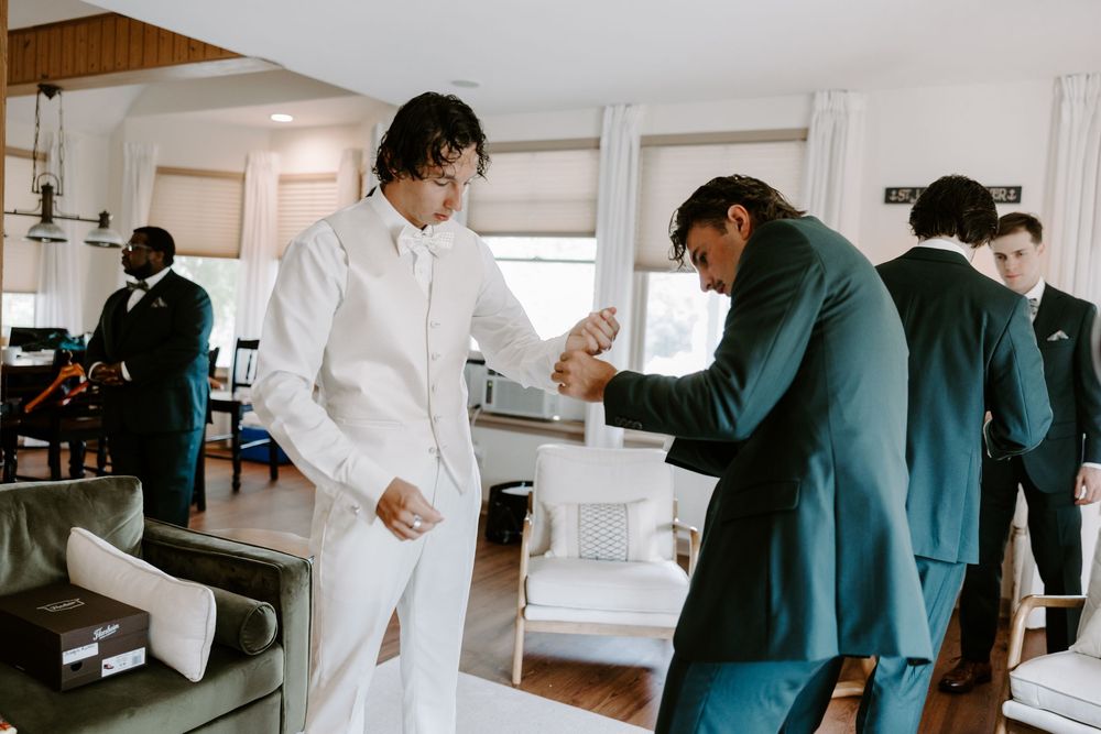 Groomsman helps groom with cufflinks as wedding party members in teal suits prepare in living room.