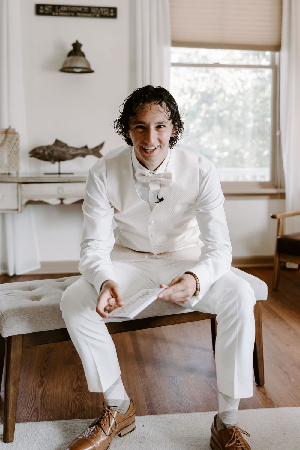 Smiling groom in white suit and bow tie sitting on bench holding wedding vows card.