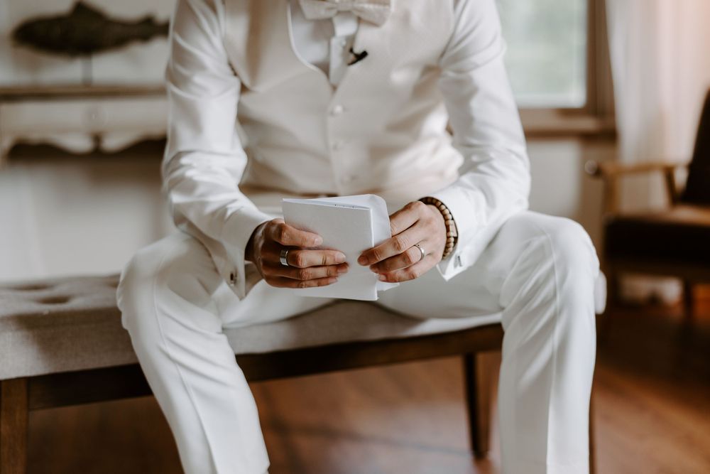 Groom in white suit and bow tie holding wedding vow card with gold bracelet detail.