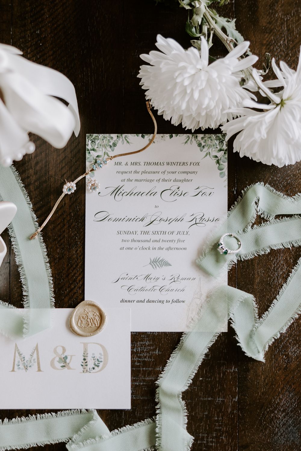 Elegant wedding invitation suite with sage green ribbons, wax seal, and white flowers.