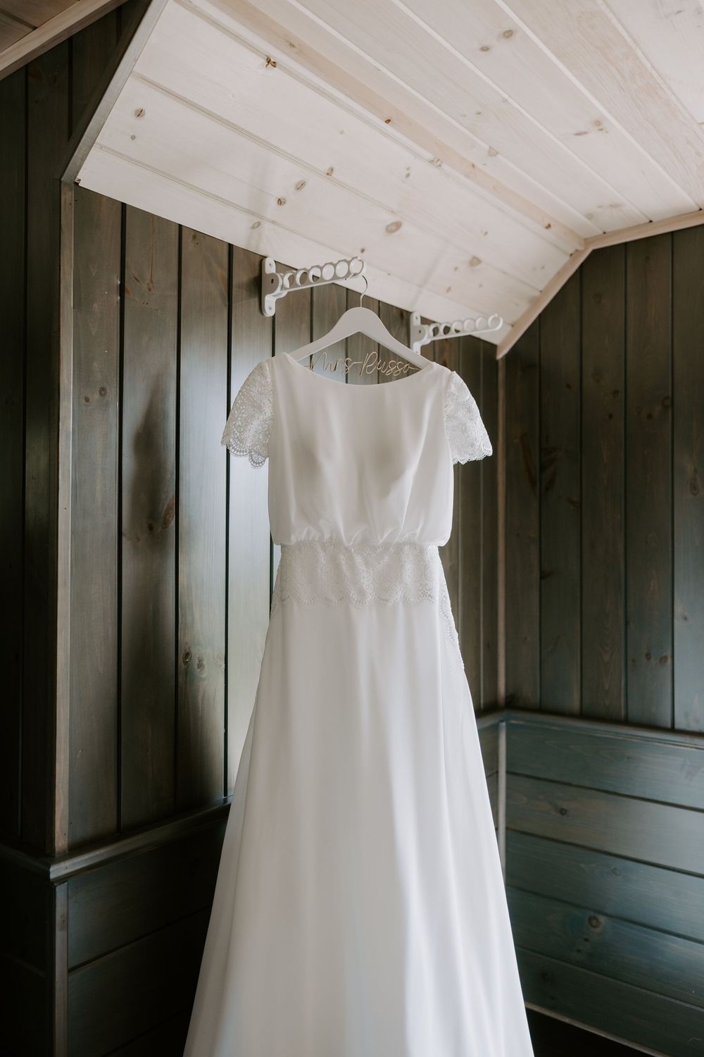 Simple white A-line wedding dress with lace cap sleeves hanging in dark wood room.