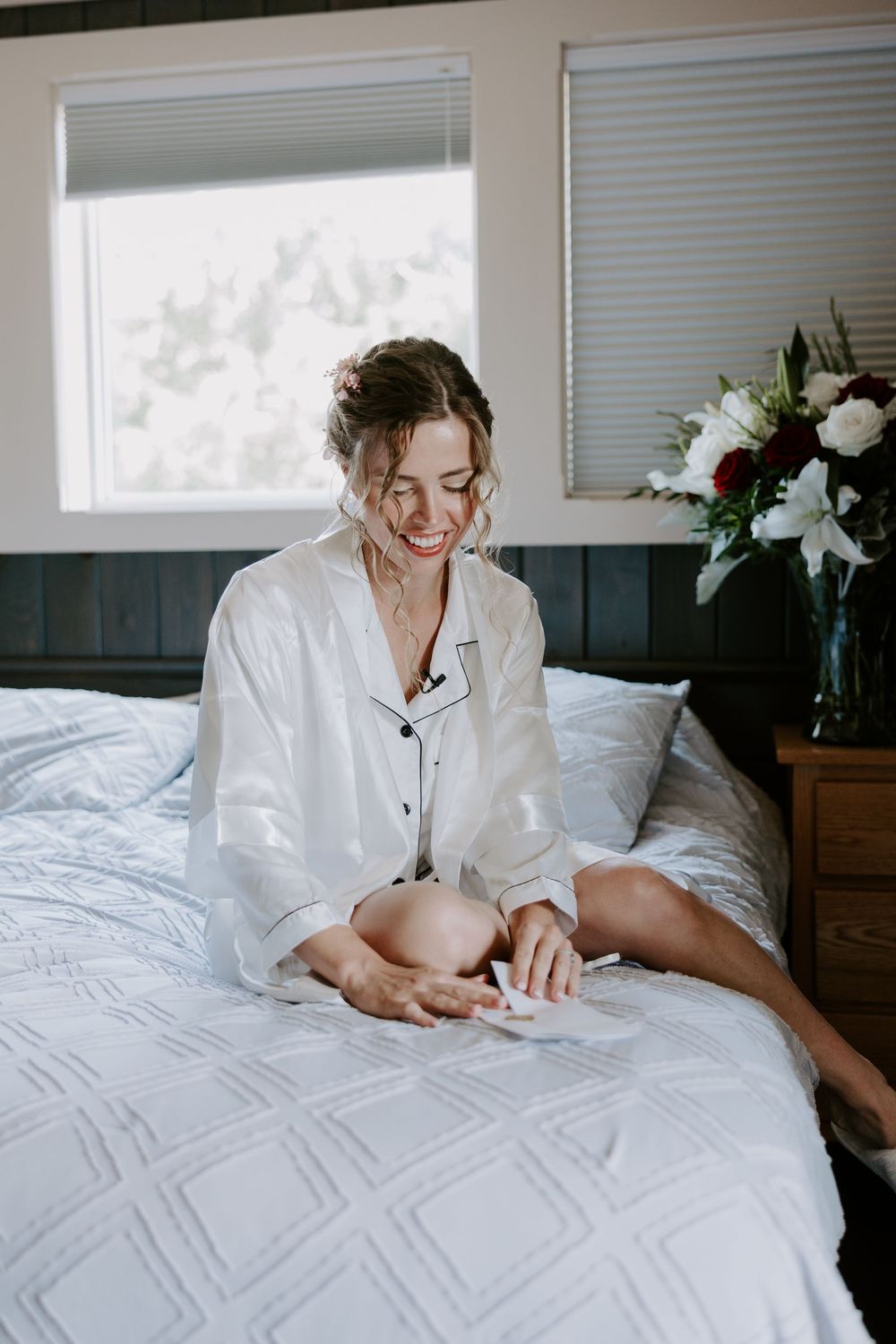 Smiling bride in white satin pajamas sits on bed reading a note with fresh flowers nearby.