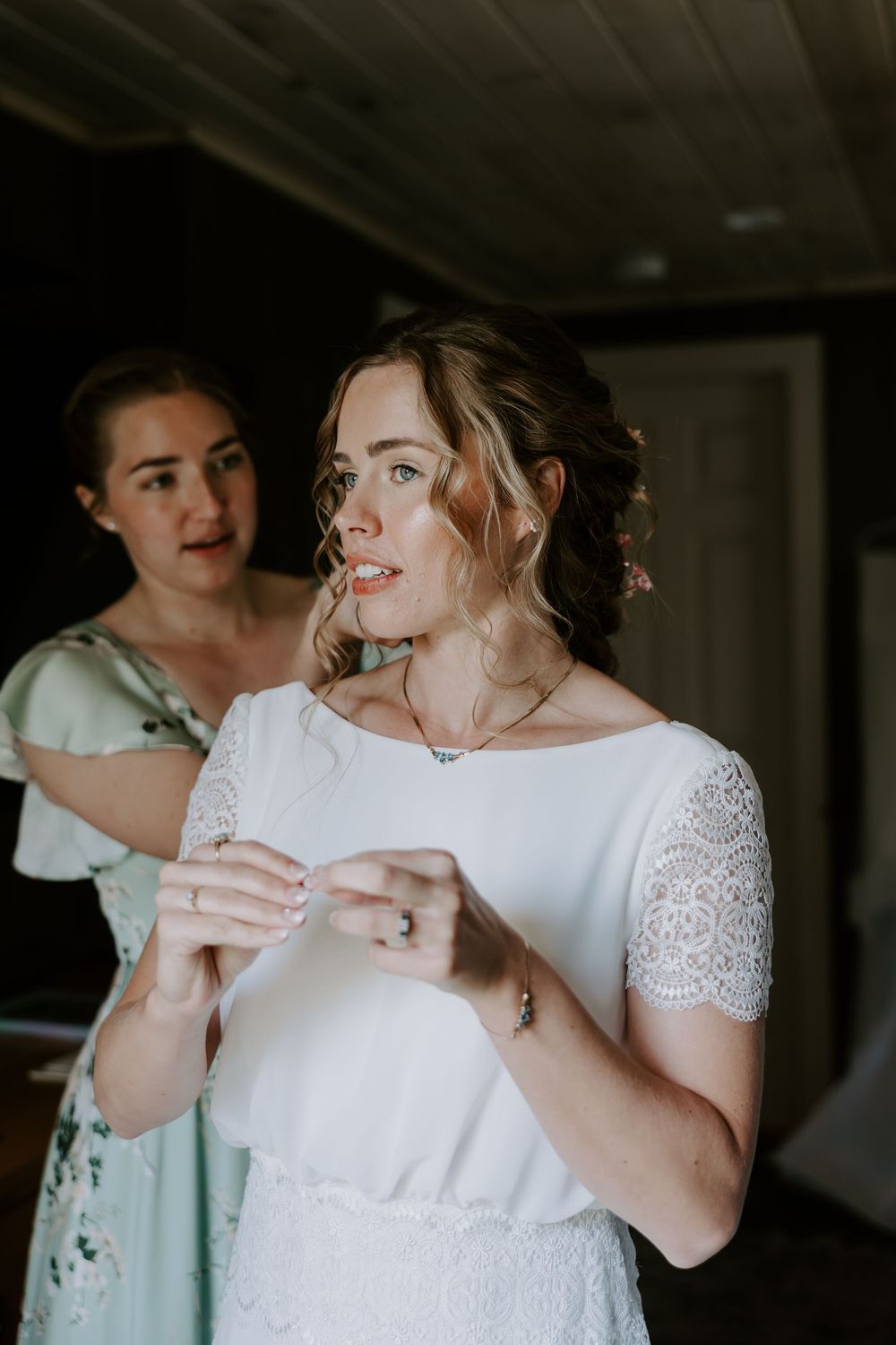 Bridesmaid helps bride into her white lace wedding dress inside a rustic wooden cabin room.