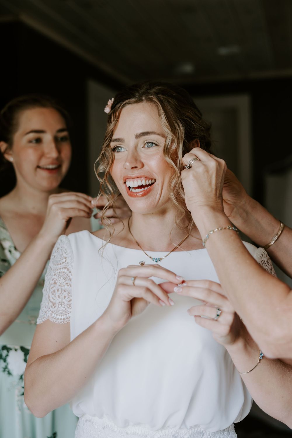 Joyful bride laughs as two women help fasten her necklace while she wears her white lace gown.