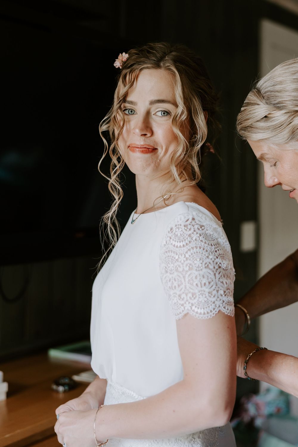 Bride in white lace wedding dress looks over shoulder as someone fastens the back of her gown.