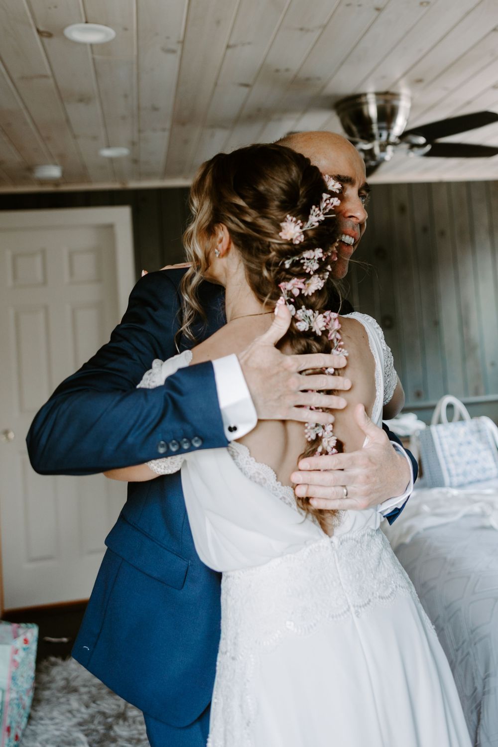 Groom in navy suit embraces bride from behind, admiring her floral hair accessories and lace gown.
