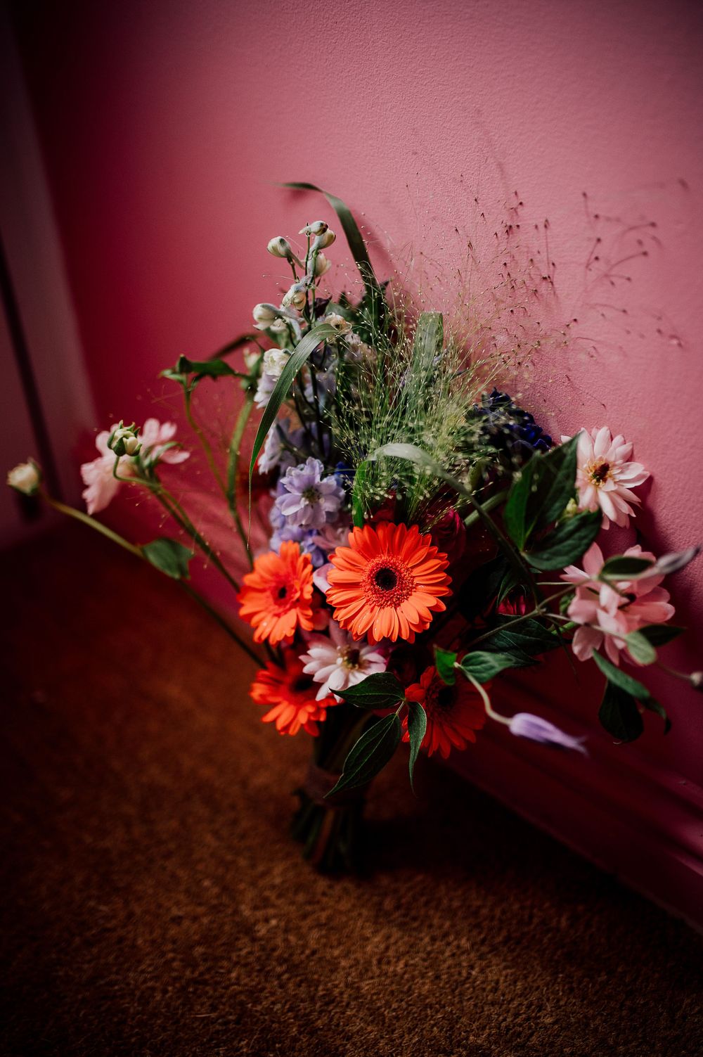 Orange gerbera daisies and white flowers arranged against a pink wall with dark brown carpet below.